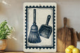 a white sign with a black and white illustration of a broom and a dustpan, placed on a wooden surface next to a wooden cutting board and a potted plant.