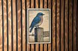 A framed illustration of a bluebird perched on a wooden post is displayed on a wooden wall.