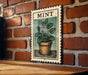 A framed illustration of a mint plant is displayed on a brick wall, with a wooden table below it.