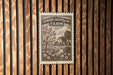 A wooden wall with a framed image of a farm scene, including a tree, house, and field, hanging on it.
