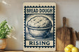 a sign that reads "BREAD DOUGH RISING" with a picture of a bowl of dough on it. The sign is placed on a wooden surface, and there are some kitchen utensils, such as a knife and a spoon, visible in the background.