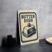 A sign that reads "BUTTER DISH" with an illustration of a butter dish on it, placed on a countertop next to some black containers.
