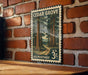 A vintage-style U.S. postage stamp depicting a scenic forest scene with tall trees and a winding path, mounted on a brick wall.