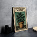 A framed illustration of a mint plant in a pot is displayed on a gray wall, accompanied by a stack of black camera lenses and a white dish.