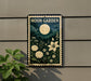 a sign for a "Moon Garden" with a night sky and flowers, mounted on a wall next to a plant.