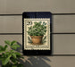 A potted plant with green leaves and a basil plant is displayed on a wall, with a 20-cent stamp featuring a culinary herb planter in the background.