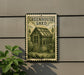 A metal sign with a picture of a greenhouse on it, mounted on a wall next to a plant.