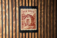 A wooden wall with a red and white postage stamp featuring a small house and a cow, mounted on the wall.