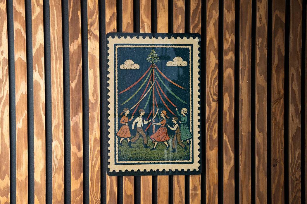 A framed illustration depicting a group of people dancing around a tall pole with colorful ribbons, set against a dark background.