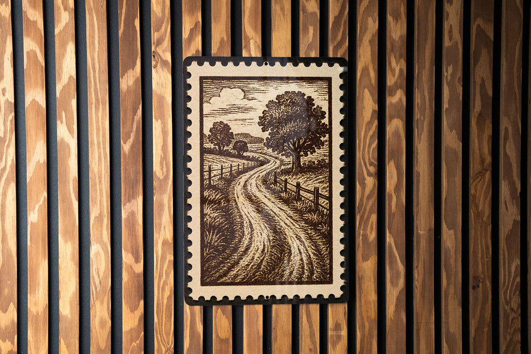a framed illustration of a rural landscape, including a winding road and trees, mounted on a wooden wall.