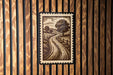 a framed illustration of a rural landscape, including a winding road and trees, mounted on a wooden wall.