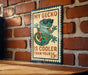 A sign featuring a cartoon gecko wearing sunglasses and stating "My gecko is cooler than your 15c" is displayed on a brick wall.
