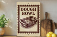 a sign that reads "DOUGH BOWL" with a picture of a dough bowl on it, placed on a shelf next to a wooden cutting board and some lemons.