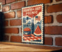 A vintage-style poster for the Fourth of July picnic, featuring a hamburger, watermelon, and an American flag, is displayed on a brick wall.