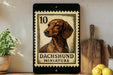 a framed stamp-like artwork depicting a dachshund miniature dog, placed on a wooden surface next to a wooden cutting board and some lemons.