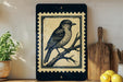 a framed illustration of a bird perched on a branch, placed on a shelf next to a wooden cutting board and some lemons.