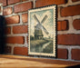 A framed illustration of a windmill is hanging on a brick wall.