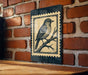 A framed illustration of a bird is hanging on a brick wall.