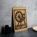 a wooden sign with a black and white illustration of a spinning wheel, placed on a gray countertop next to a stack of black camera lenses.