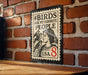 A metal sign with a stamp-like design hangs on a brick wall, featuring a woman with birds perched on her shoulders.