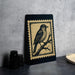 a framed illustration of a bird perched on a branch, placed on a table next to some black camera lenses.