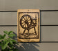 a wooden spinning wheel on a wall, with a plant to the left of the frame.