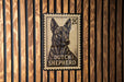 A postage stamp featuring a dog, specifically a German Shepherd, is displayed on a wooden wall.