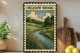 a framed illustration of a scenic landscape with a river, flowers, and trees, displayed on a shelf next to a wooden cutting board and a potted plant.