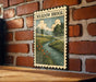 a framed illustration of a countryside landscape with a river and flowers, mounted on a brick wall.
