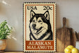 A metal sign featuring a black and white illustration of a husky dog, with text indicating it is a 20 cent United States postage stamp for Alaska Malamute dogs.