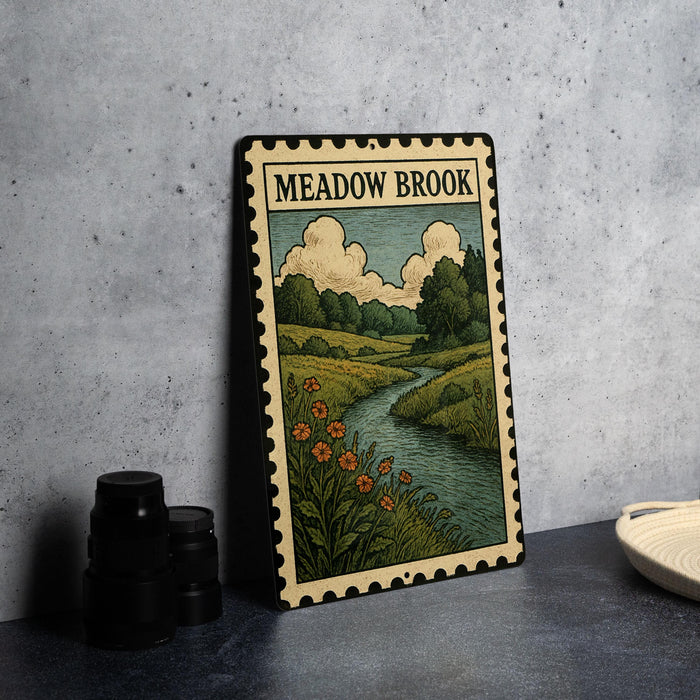 a framed illustration of a scenic landscape with a river, flowers, and trees, placed on a table next to some black camera lenses.