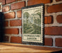 A metal sign with a stamp-like design hangs on a brick wall, depicting a garden scene with a greenhouse and plants.