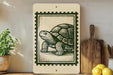 a wooden cutting board with a decorative stamp of a turtle on it, placed on a shelf next to a potted plant and some lemons.