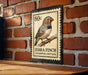 A metal postage stamp featuring a detailed illustration of a zebra finch perched on a branch, with the text "Zebra Finch" and "Tæeniopygia gutta" printed on it.