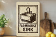 a sign that reads "FARMHOUSE SINK" with a black and white illustration of a sink. The sign is placed on a wooden surface, and there are some kitchen utensils and a potted plant nearby.