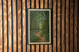 a framed painting of a forest path, hanging on a wooden wall.