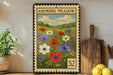 a framed print of a colorful floral scene, with a postage stamp-like border, placed on a wooden surface next to a wooden cutting board and some potted plants.