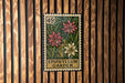 A metal stamp with a floral design, featuring two pink flowers and a yellow flower, is displayed on a wooden wall.