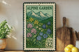 A framed postage stamp depicting a mountain landscape with colorful flowers, placed on a shelf next to a wooden cutting board and some potted plants.