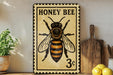 A vintage-style metal sign with a bee illustration, placed on a wooden shelf next to a potted plant and kitchen utensils.