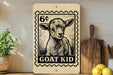 A wooden sign featuring a black and white illustration of a goat, with the text "GOAT KID" printed on it, is displayed on a shelf next to a potted plant and a wooden cutting board.