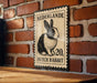 A metal sign with a black and white illustration of a Dutch rabbit, mounted on a brick wall.