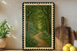 a framed painting of a forest path with a winding trail, surrounded by lush greenery and a wooden cutting board with a knife on the right side.