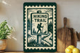 a green and white sign that reads "HIKING TRAIL" with a silhouette of a hiker, placed on a wooden surface next to a wooden cutting board and some potted plants.