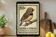 A framed postage stamp featuring a detailed illustration of a zebra finch, with the text "ZEBRA FINCH TAENIOPYGA GUTTATA" printed on it. The stamp is placed on a shelf next to a wooden cutting board and a potted plant.
