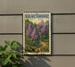 A postage stamp featuring a lush garden scene with purple flowers, mounted on a wall next to a plant.
