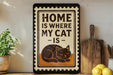 A framed sign with the text "HOME IS WHERE MY CAT IS" and an illustration of a cat is displayed on a shelf next to a wooden cutting board and a potted plant.