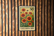 A postage stamp from Gaillardia Bed, featuring a vibrant illustration of a field of blooming flowers, is displayed on a wooden wall.