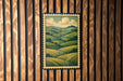a framed painting of a countryside landscape, hanging on a wooden wall.