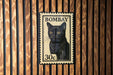 A black cat stamp with the text "BOMBAY 30c" is displayed on a wooden wall.